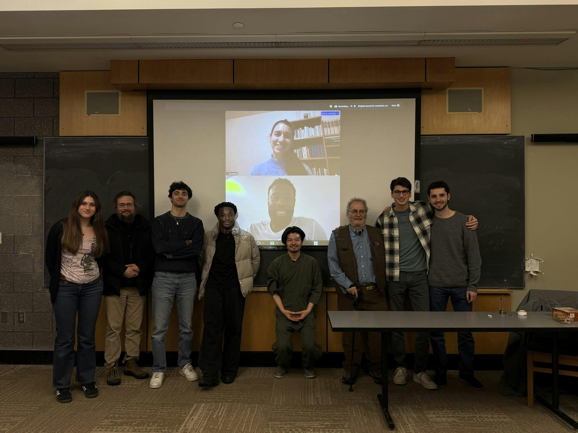 An image of students and a professor in a classroom with two students on Zoom pictured in the background on a projected screen.