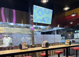 A TV with the restaurant's menu hangs above of an empty table. Behind the TV and a glass wall, a chef stands in front of a wall decorated with fried egg clipart.