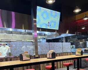 A TV with the restaurant's menu hangs above of an empty table. Behind the TV and a glass wall, a chef stands in front of a wall decorated with fried egg clipart.