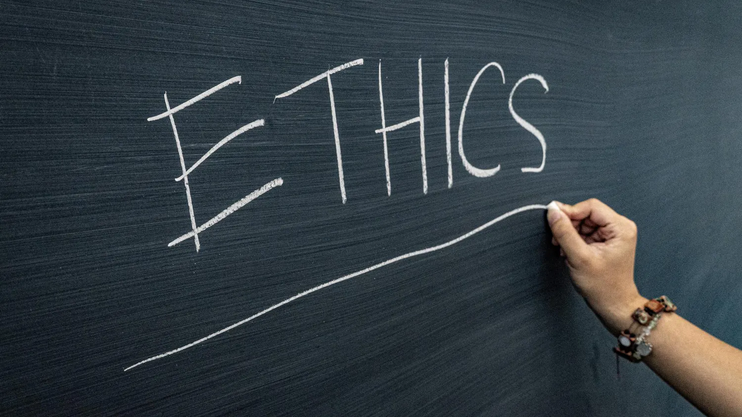 A photo of a hand drawing a line with chalk under the word “ETHICS” written on a dark green chalkboard.