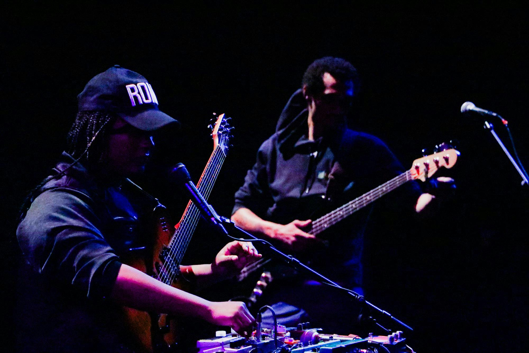 A guitarist playing in the background and a guitarist wearing a black cap playing in the front. Setting is very dark with navy hues. 
