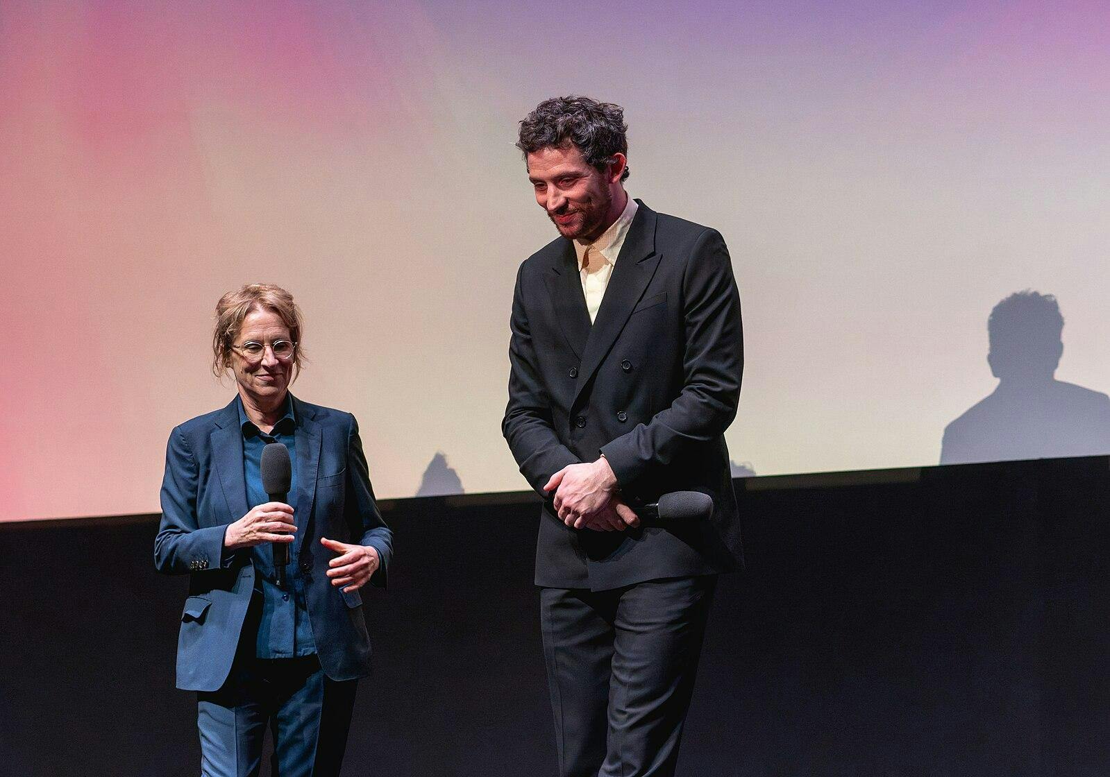 Josh O'Connor mid-bow on stage with Kelly Reichardt behind him. Both are wearing suits and holding mics.