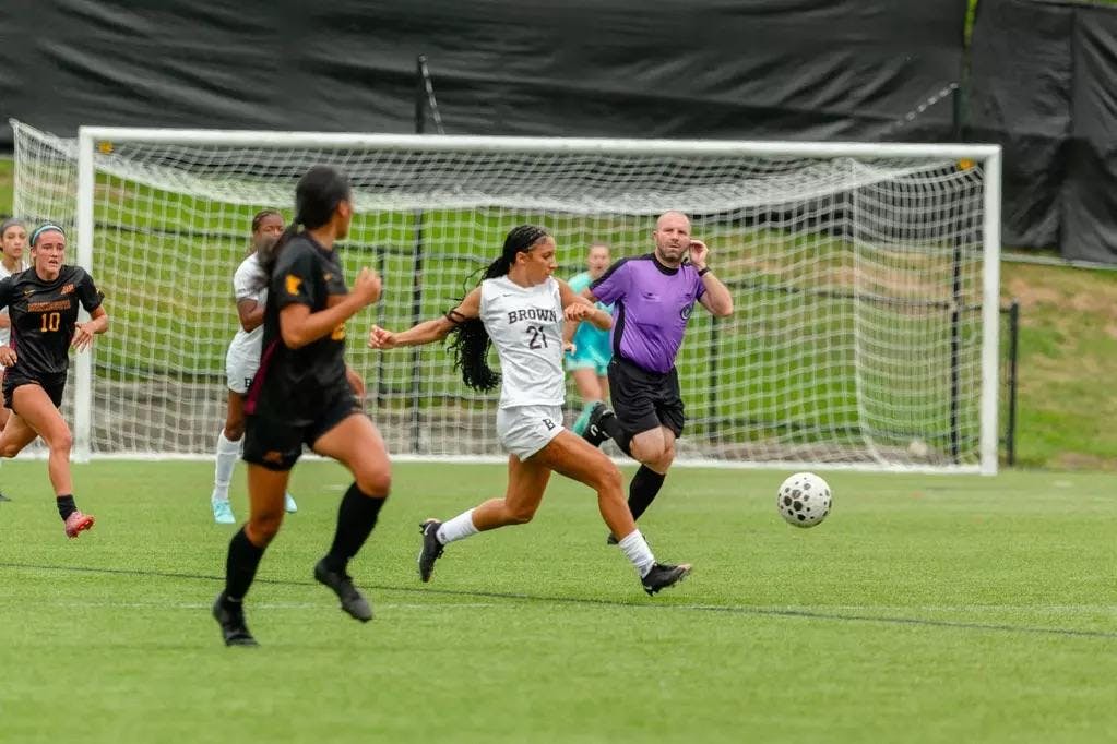A photo of a female soccer player in a white Brown University uniform running right to kick a soccer ball, with multiple players behind her and a referee dressed in purple to her left.