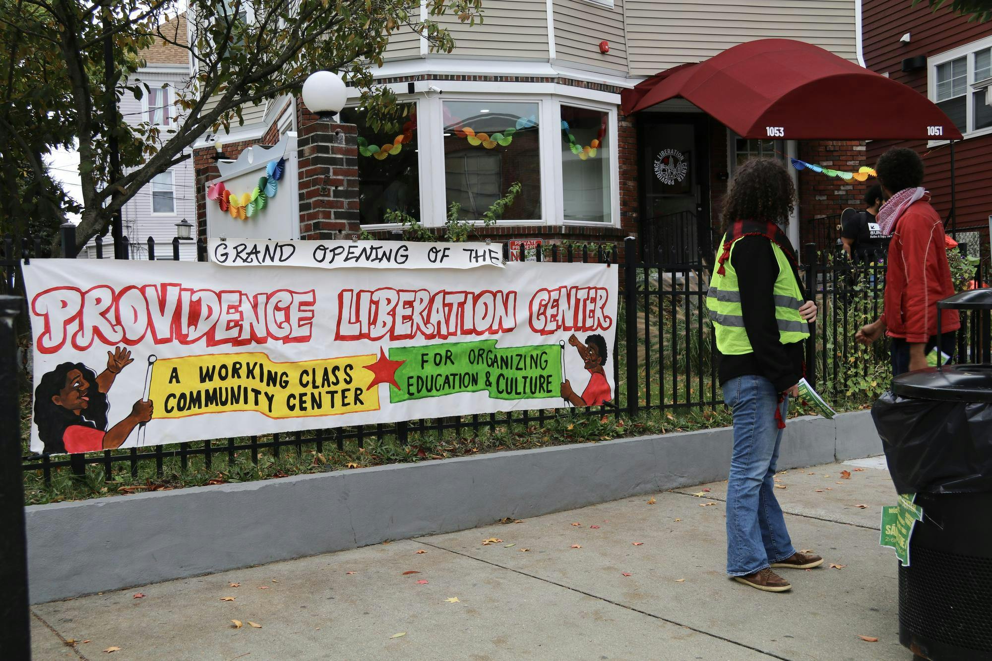 Photo of a white banner clipped onto a black fence in front of a tan house with two people standing on the right. The banner reads “Grand Opening of the Providence Liberation Center” with the text “A working class community center for organizing education & culture” underneath it on drawn yellow and green flags. 
