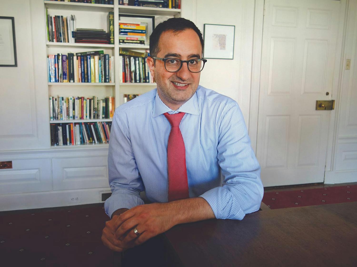 Rashid Zia, wearing glasses, a blue pinstripe shirt and a red tie, smiles at the camera in front of a wall lined with books.