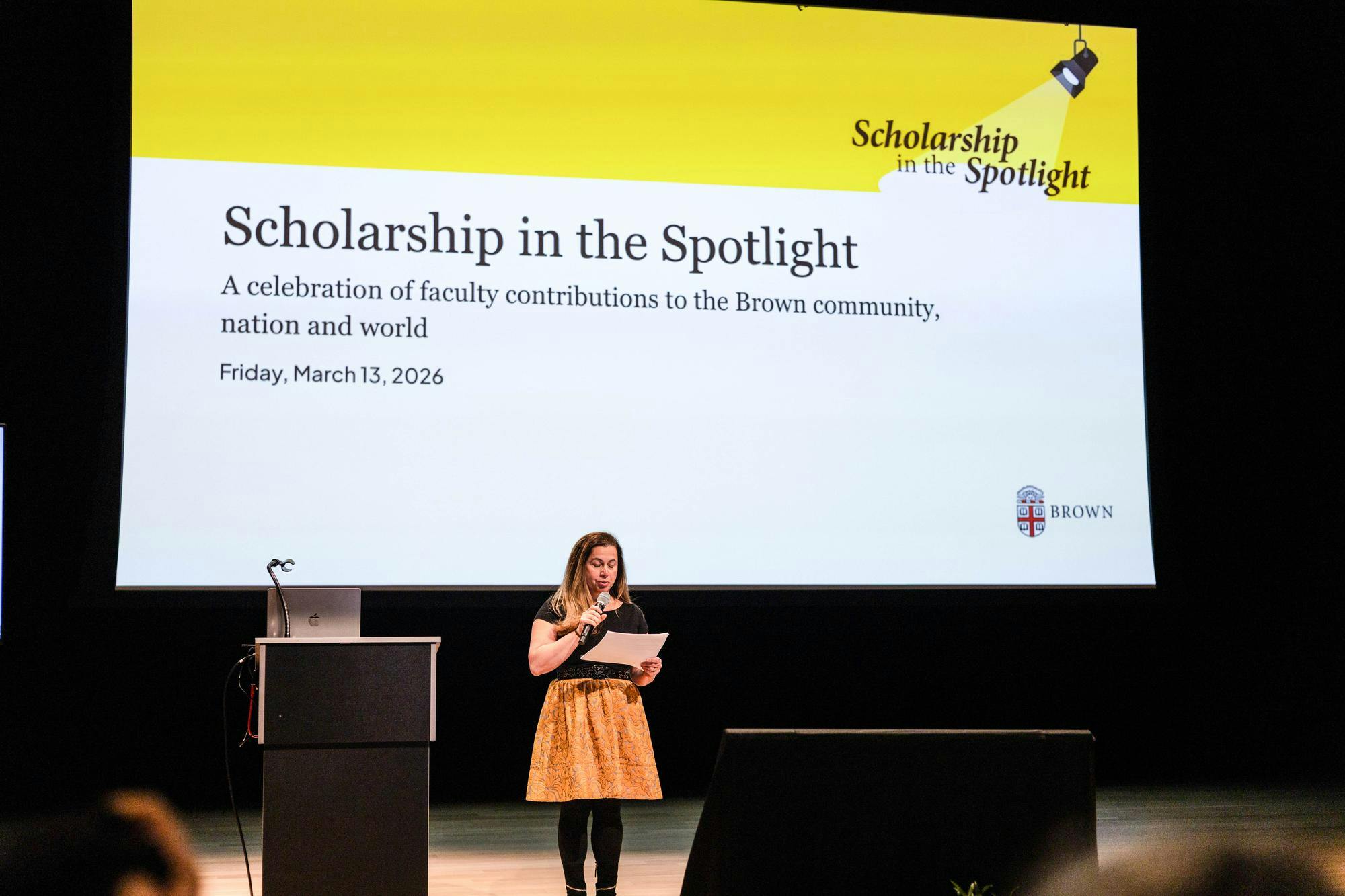 A photo of a woman speaking on a stage in front of a big screen. 

