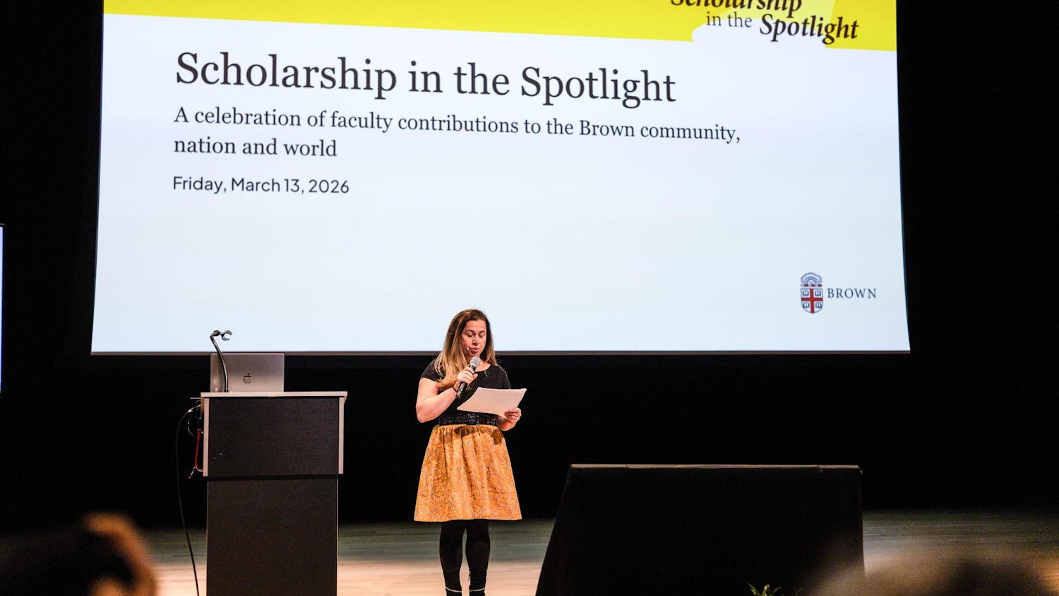 A photo of a woman speaking on a stage in front of a big screen.