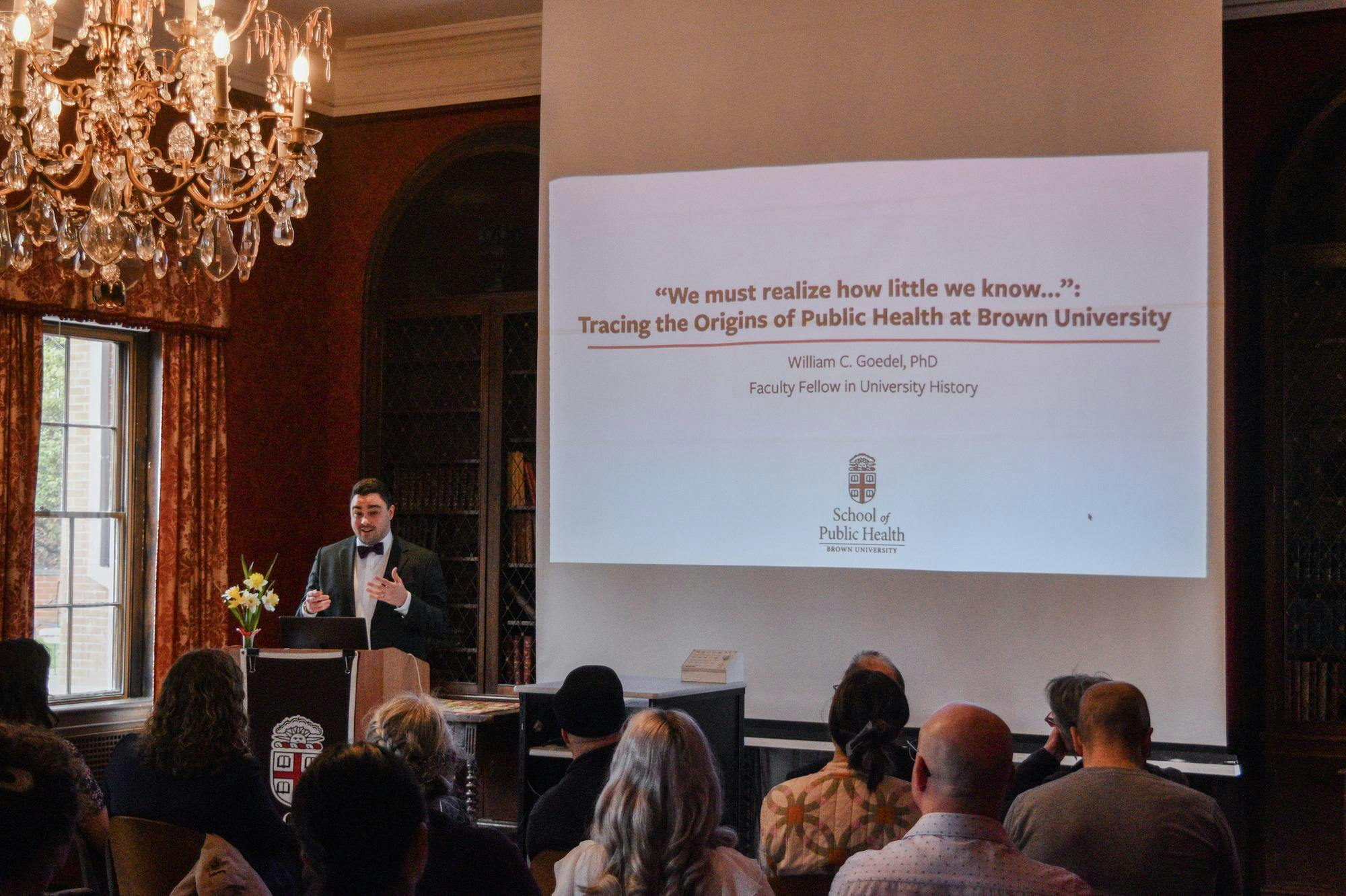 A photo showing Professor William Goedel presenting a slideshow on Public Health at Brown University in front of an audience in a room with a chandelier. 