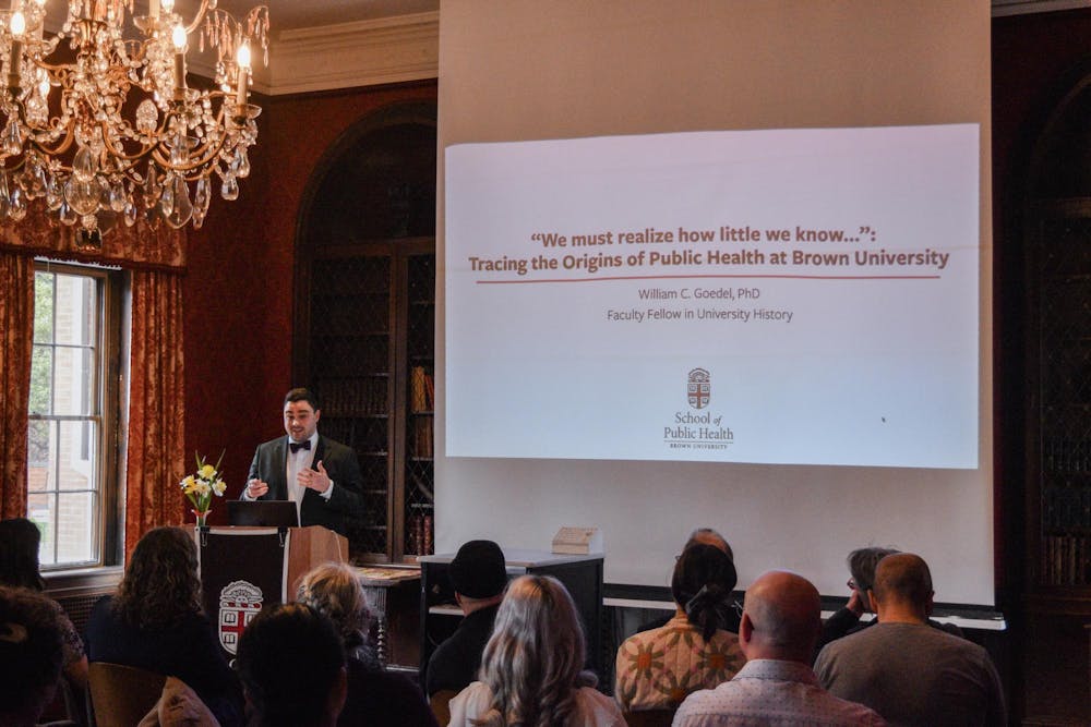 A photo showing Professor William Goedel presenting a slideshow on Public Health at Brown University in front of an audience in a room with a chandelier. 