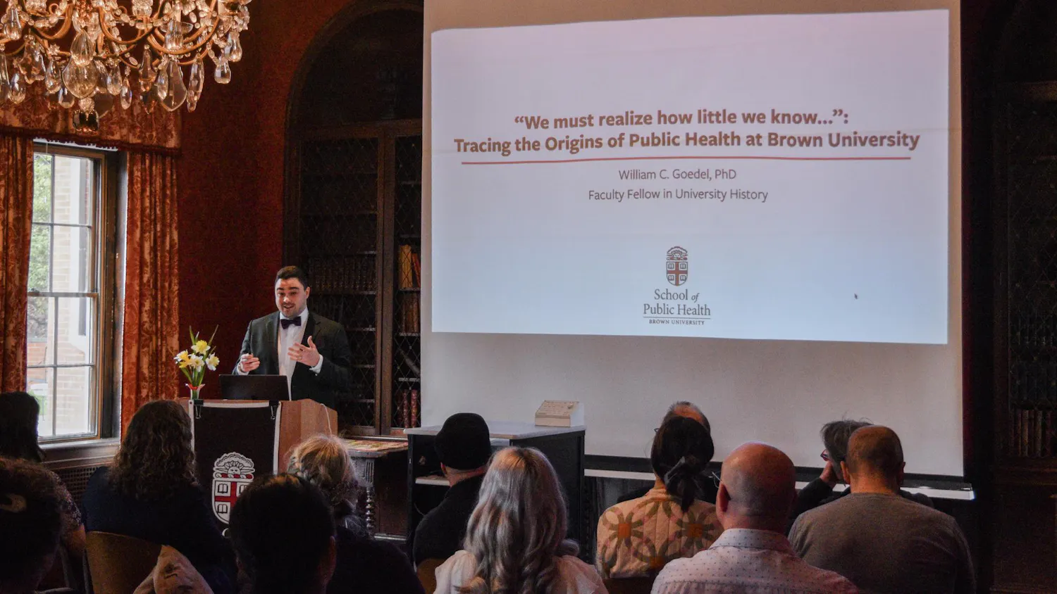 A photo showing Professor William Goedel presenting a slideshow on Public Health at Brown University in front of an audience in a room with a chandelier.
