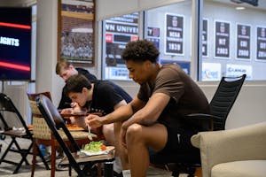 Lewis sits in an office chair and extends a fork toward a plate of salad. Two teammates sit behind him, also eating.