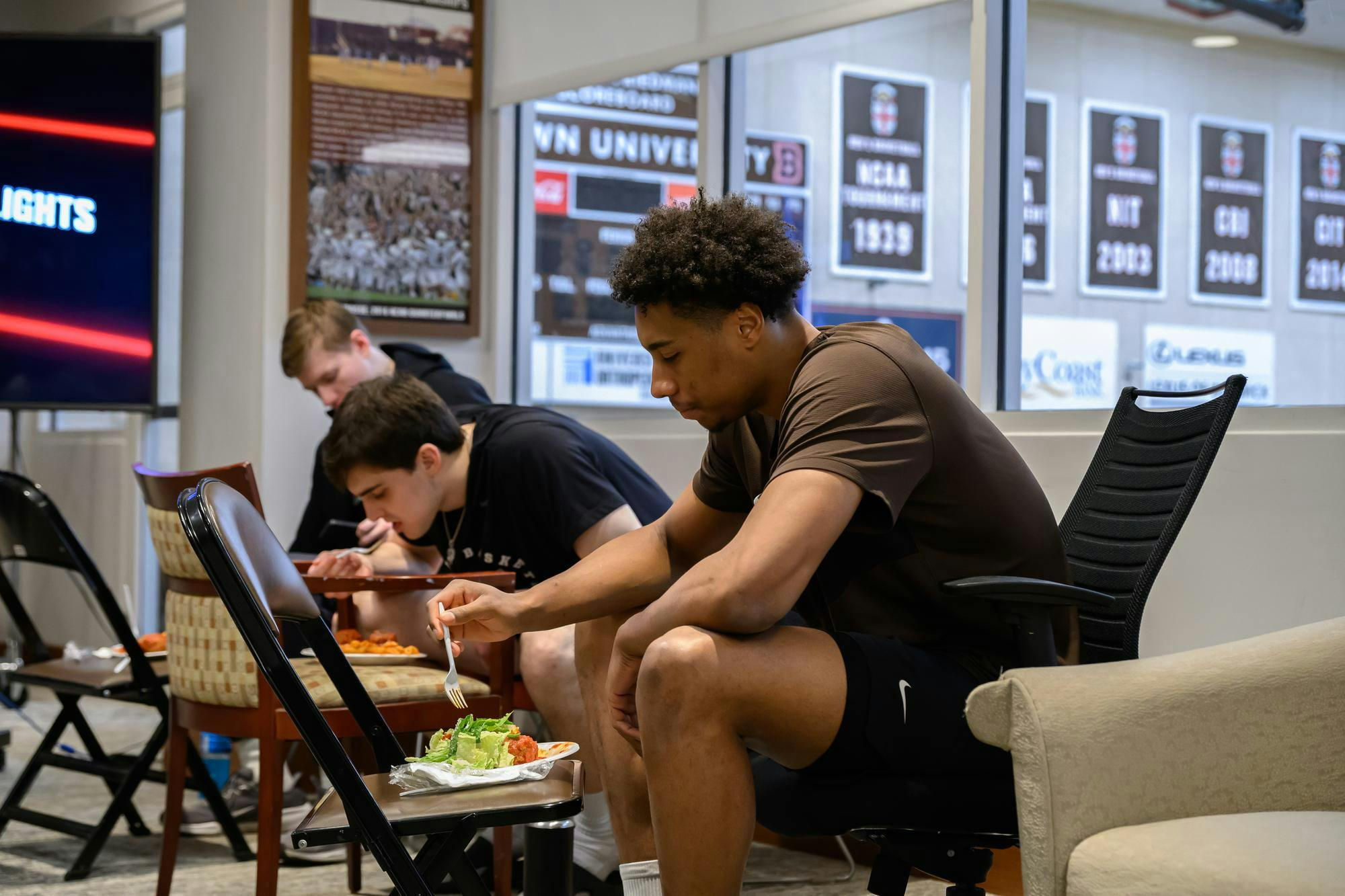 Lewis sits in an office chair and extends a fork toward a plate of salad. Two teammates sit behind him, also eating.