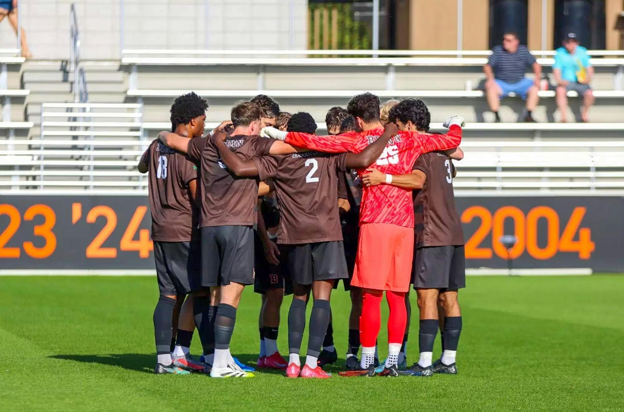 A soccer team of students wearing brown jerseys and the goalie wearing a red jersey all huddling on a field. A white number 2 is visible on the back of one student's jersey.