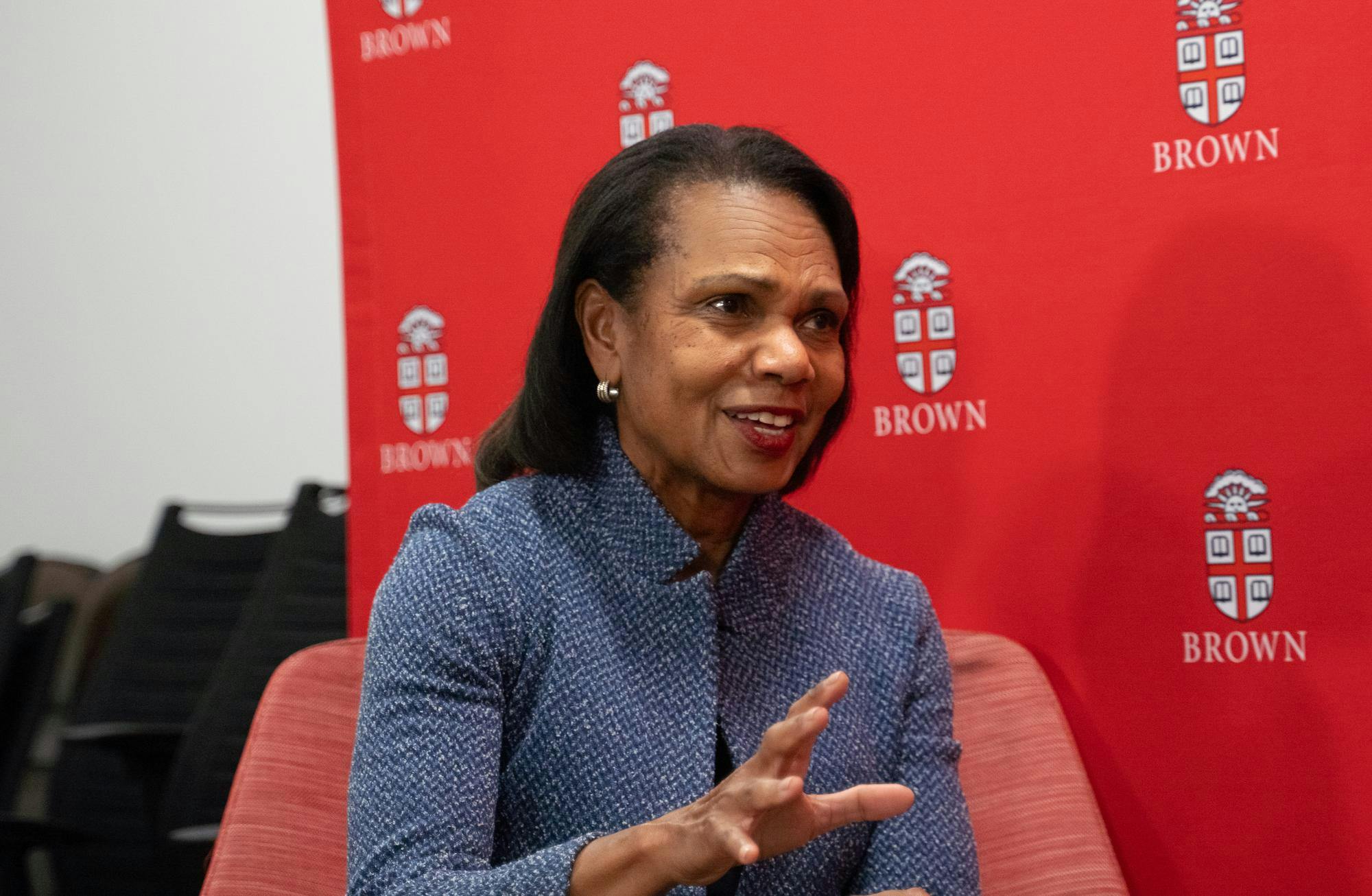 Photo of of Condoleezza Rice gesturing to someone to the right of the viewer, with a red banner patterned Brown's logo behind her