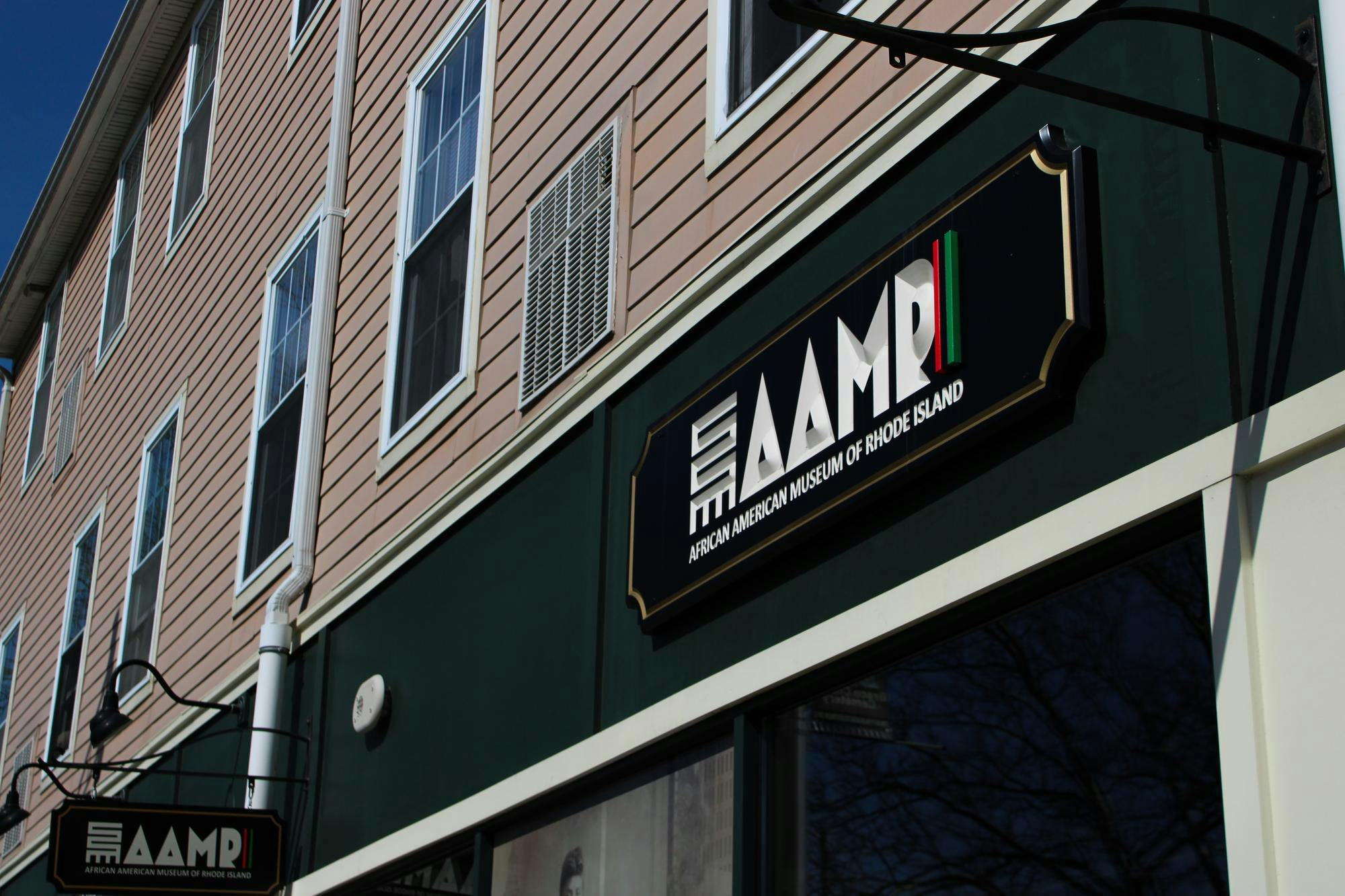 Photo of a sign on a green strip on a building saying “AAMRI” in all capital letters. The I is a strip with a red, black and green stripe. Below, “African American Museum of Rhode Island” is written in white text and all capital letters. All on a black plaque.