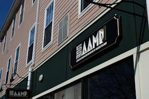 Photo of a sign on a green strip on a building saying “AAMRI” in all capital letters. The I is a strip with a red, black and green stripe. Below, “African American Museum of Rhode Island” is written in white text and all capital letters. All on a black plaque.