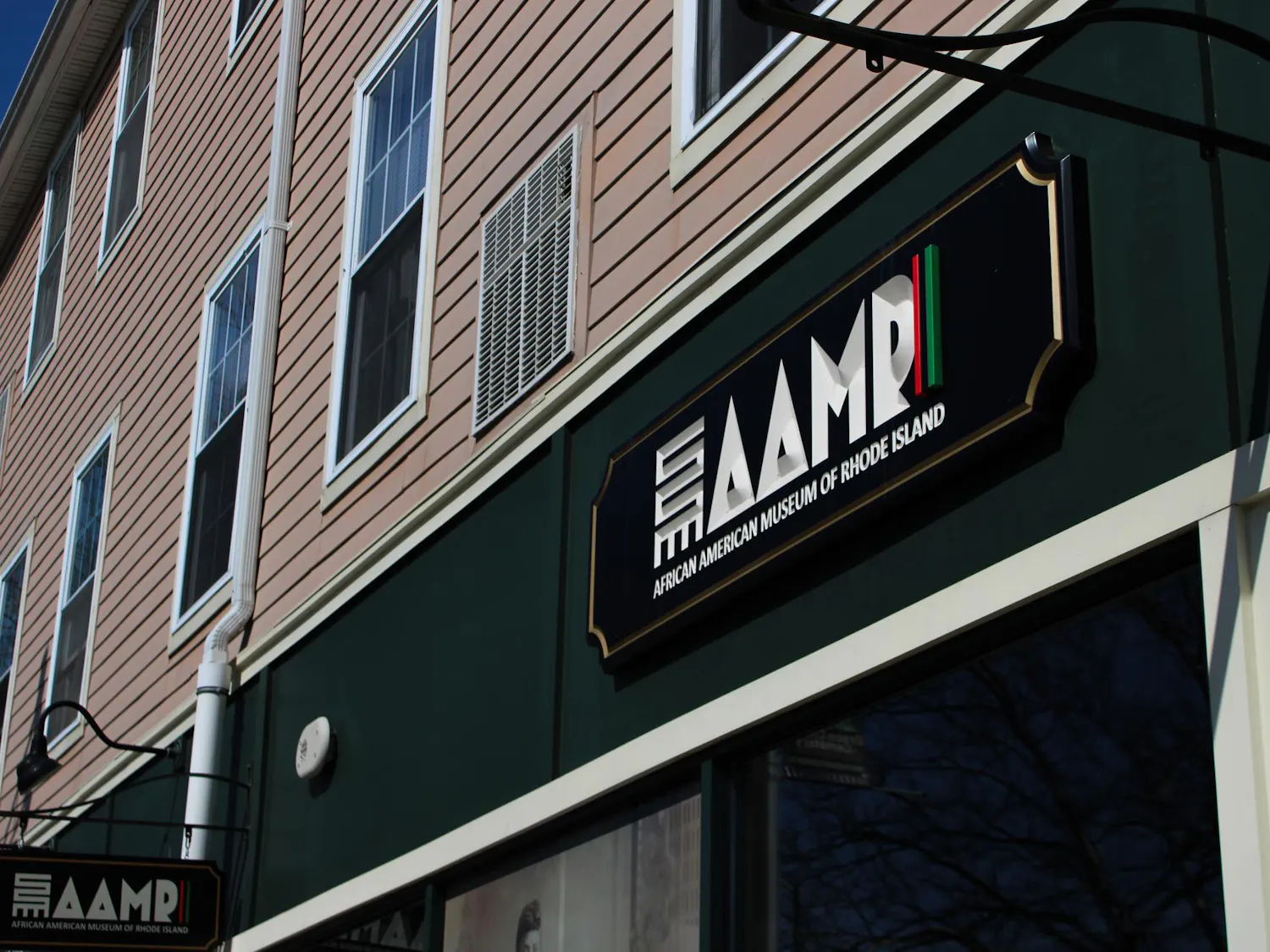 Photo of a sign on a green strip on a building saying “AAMRI” in all capital letters. The I is a strip with a red, black and green stripe. Below, “African American Museum of Rhode Island” is written in white text and all capital letters. All on a black plaque.