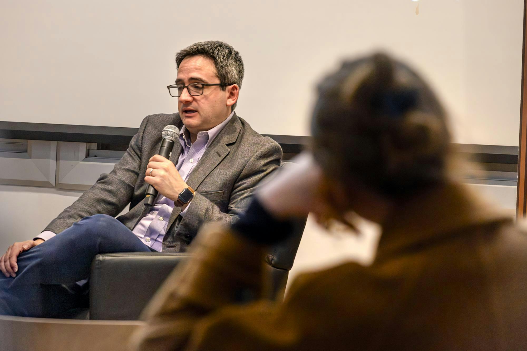 Image of Oren Cass in a blue shirt and gray blazer sitting with his legs crossed and holding a microphone, with a blurred figure of a woman resting her head on her arm in the foreground. 
