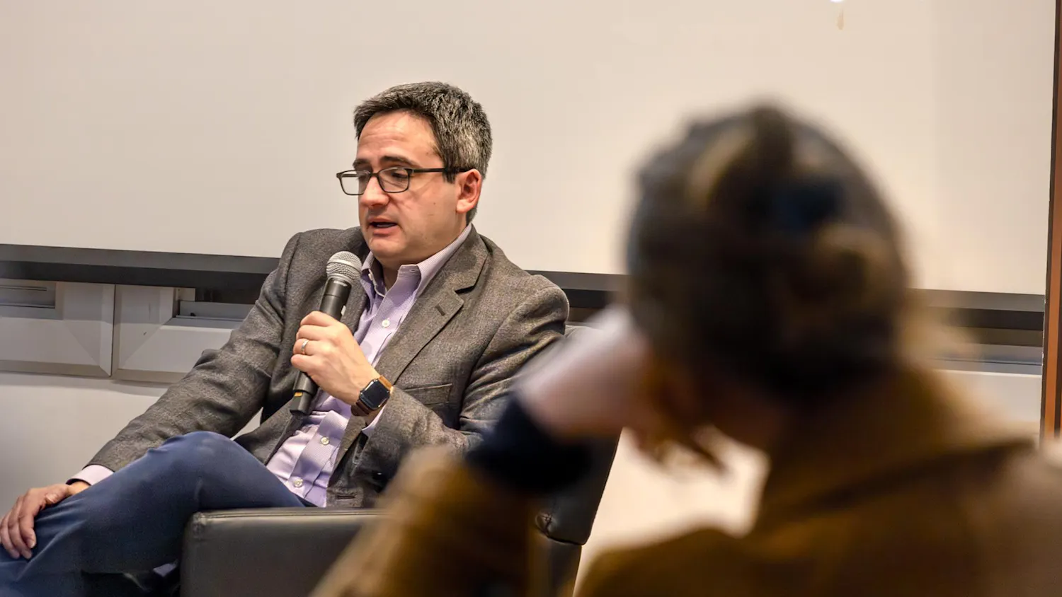 Image of Oren Cass in a blue shirt and gray blazer sitting with his legs crossed and holding a microphone, with a blurred figure of a woman resting her head on her arm in the foreground.