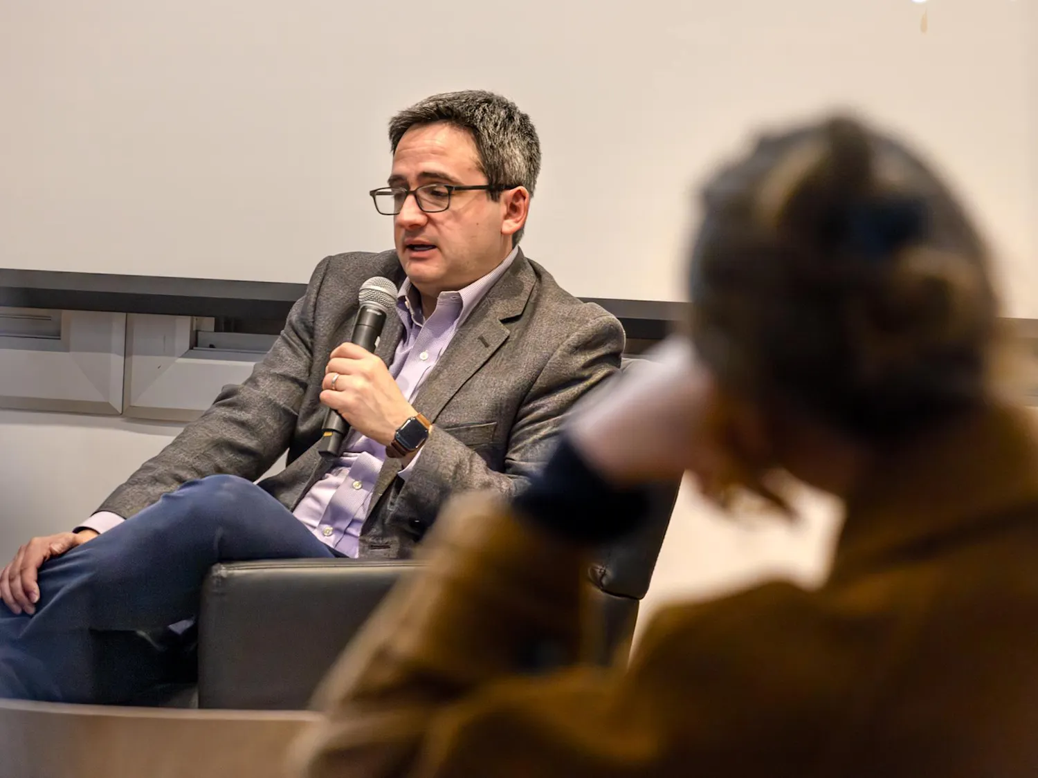 Image of Oren Cass in a blue shirt and gray blazer sitting with his legs crossed and holding a microphone, with a blurred figure of a woman resting her head on her arm in the foreground.