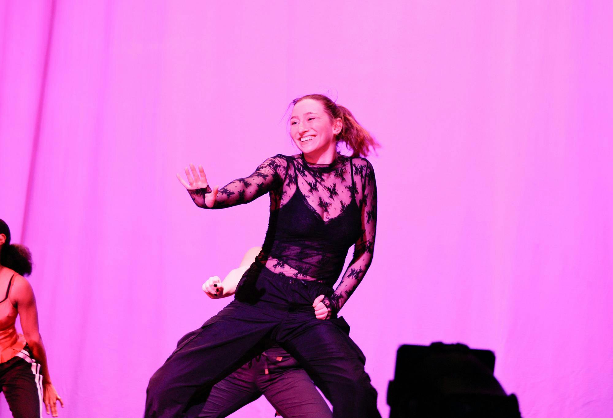 A dancer in front of a pink background extending one arm out with palm facing the audience.