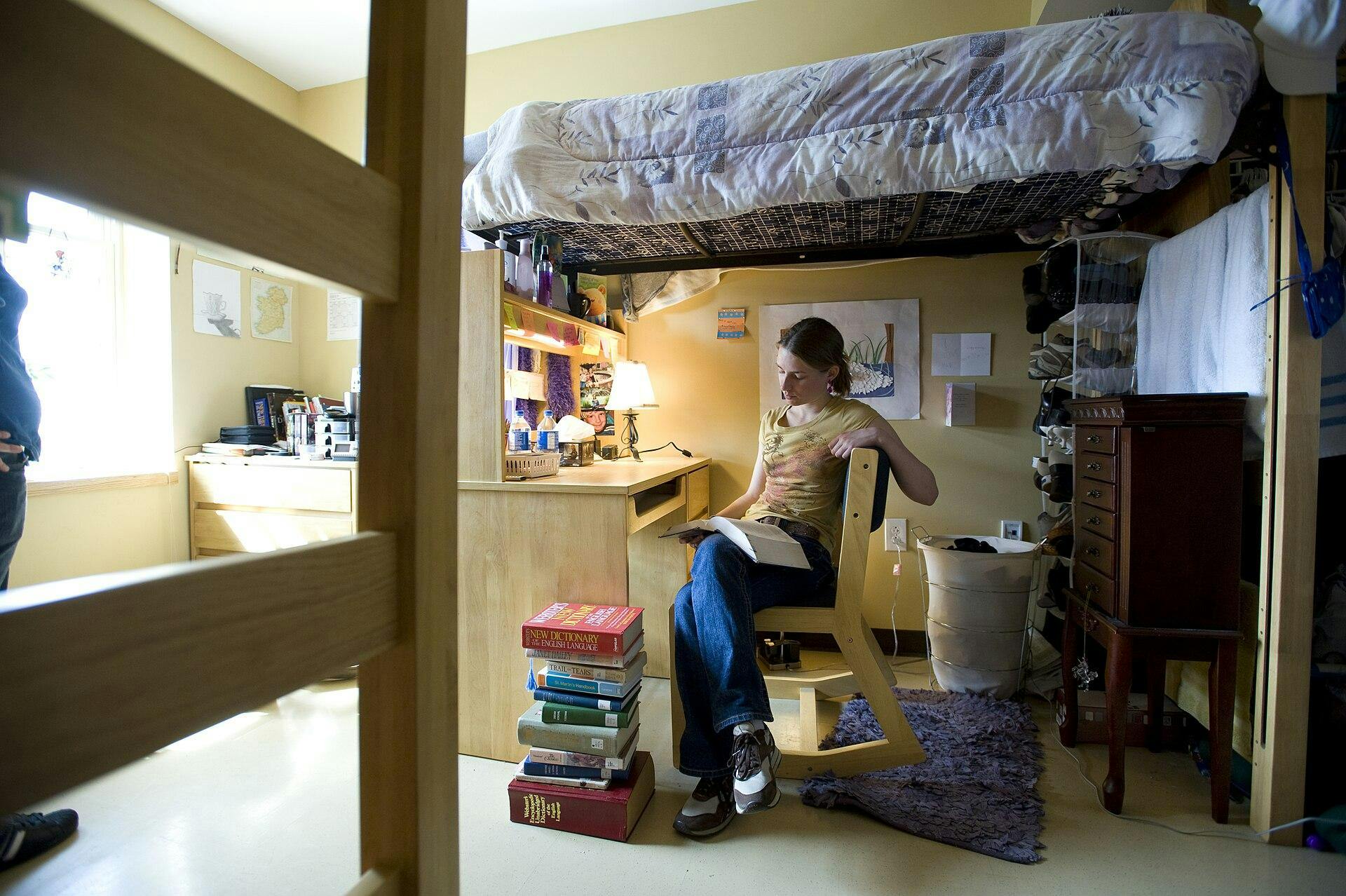 A woman sits in a dorm room under a lofted bed. 
