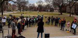 A speaker stands on the steps of the campus center, overlooking the Main Green. A crowd of about 30 people stand listening.