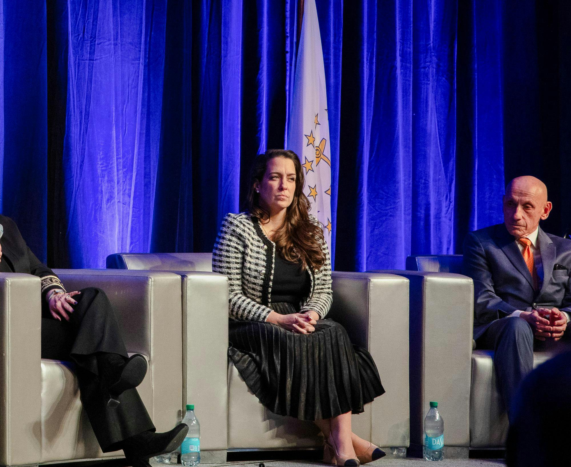 A photo of a lady sitting on a couch in front of a flag, with two people to the left and right of her.