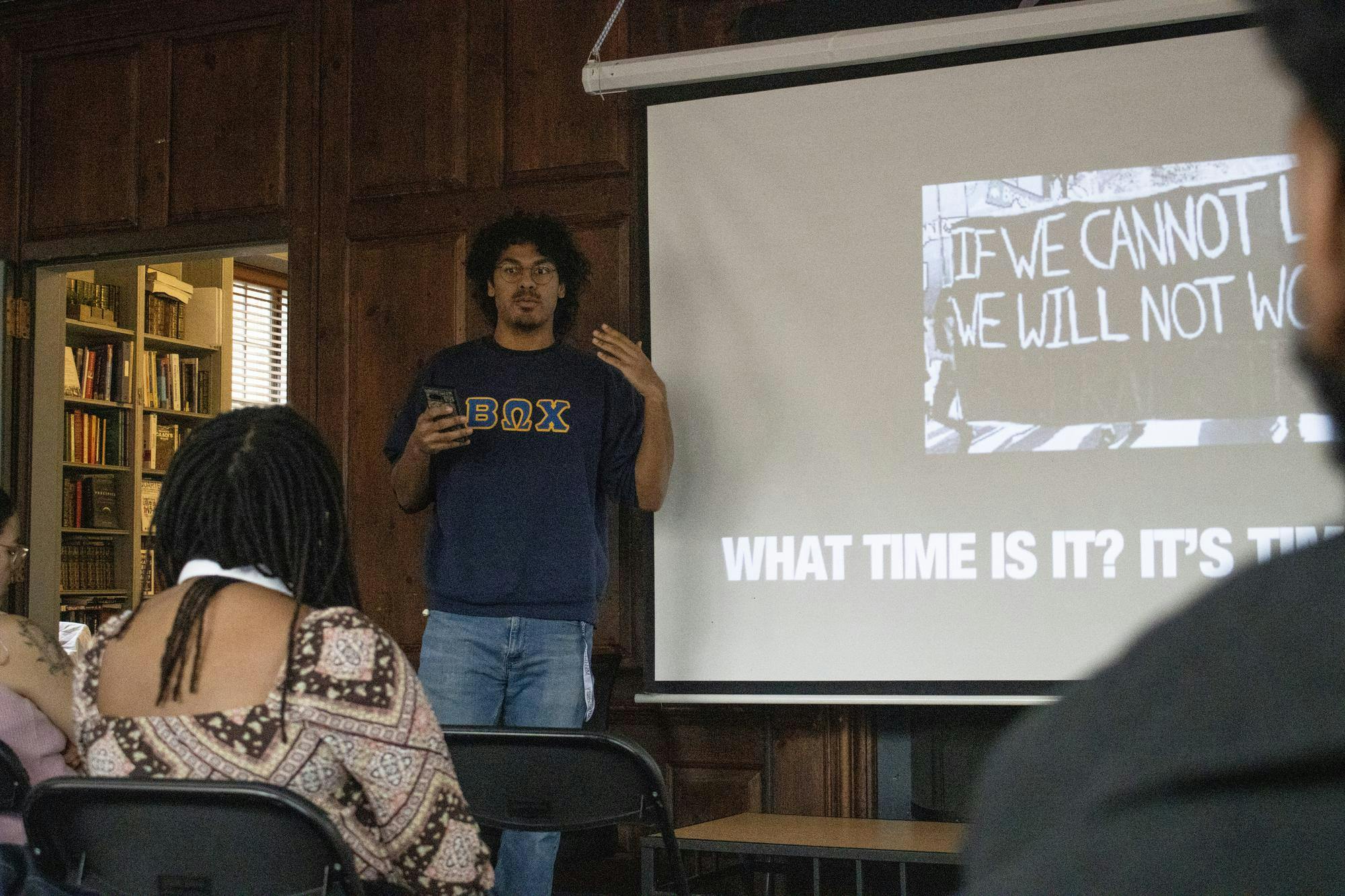 A member of the Beta Omega Chi fraternity is standing in the front of the room speaking. There is a slide behind him that reads “What time is it?”