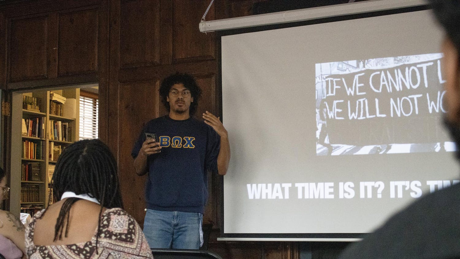 A member of the Beta Omega Chi fraternity is standing in the front of the room speaking. There is a slide behind him that reads “What time is it?”