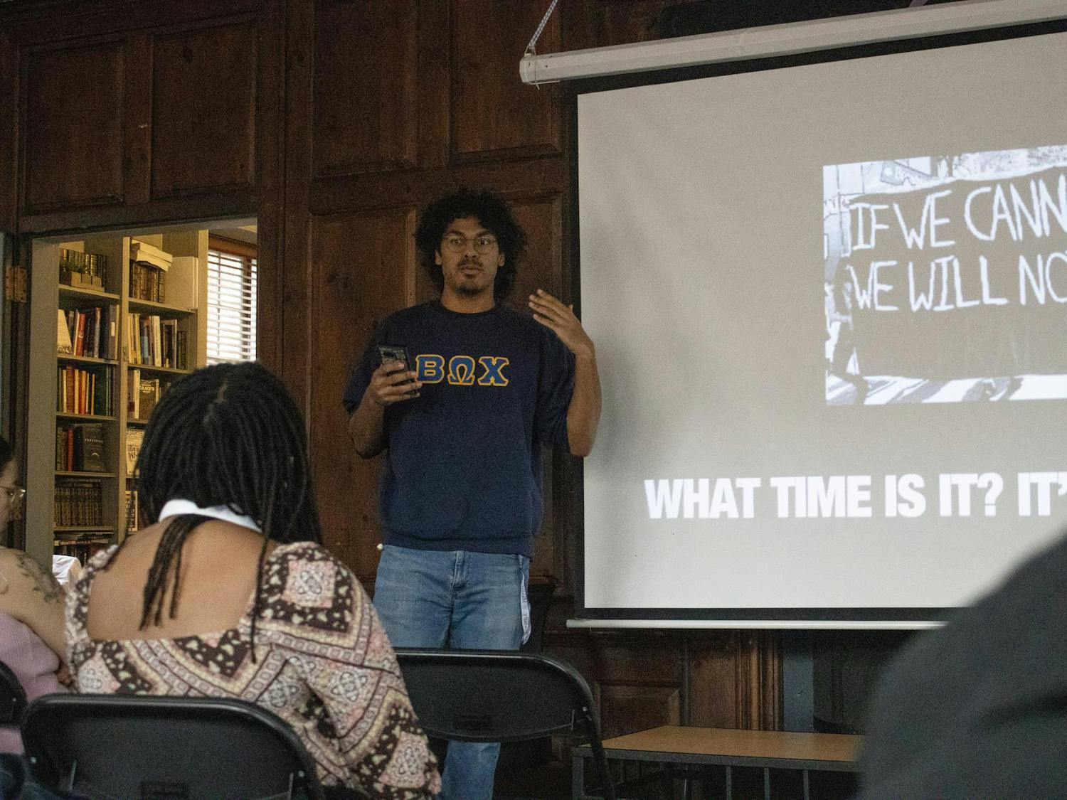 A member of the Beta Omega Chi fraternity is standing in the front of the room speaking. There is a slide behind him that reads “What time is it?”
