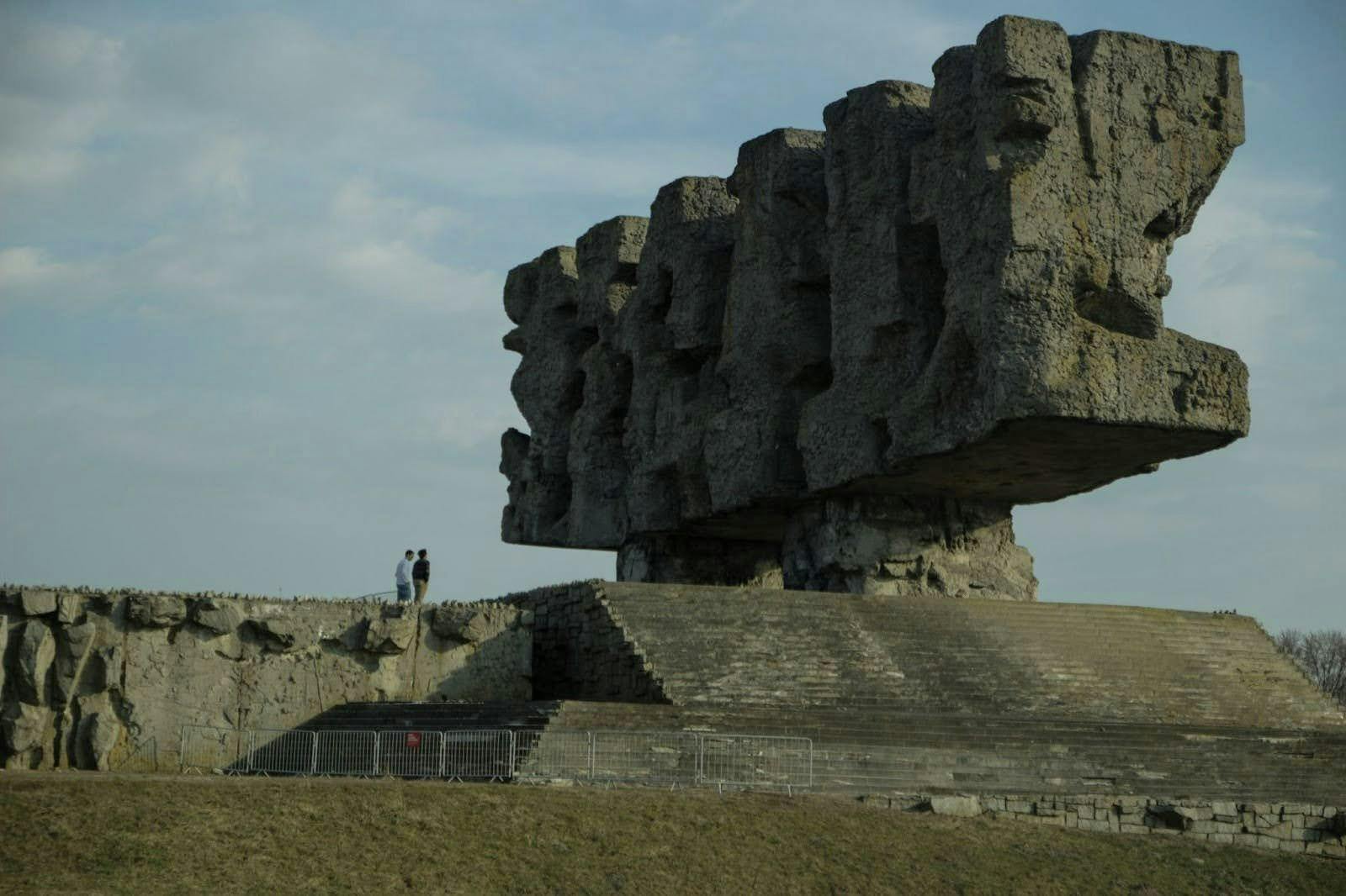 A photo showing a large, rectangular stone sculpture outside with stairs leading up to its platform. Two people are standing on the platform next to the sculpture. 