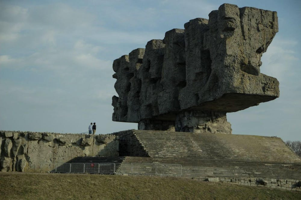 A photo showing a large, rectangular stone sculpture outside with stairs leading up to its platform. Two people are standing on the platform next to the sculpture. 