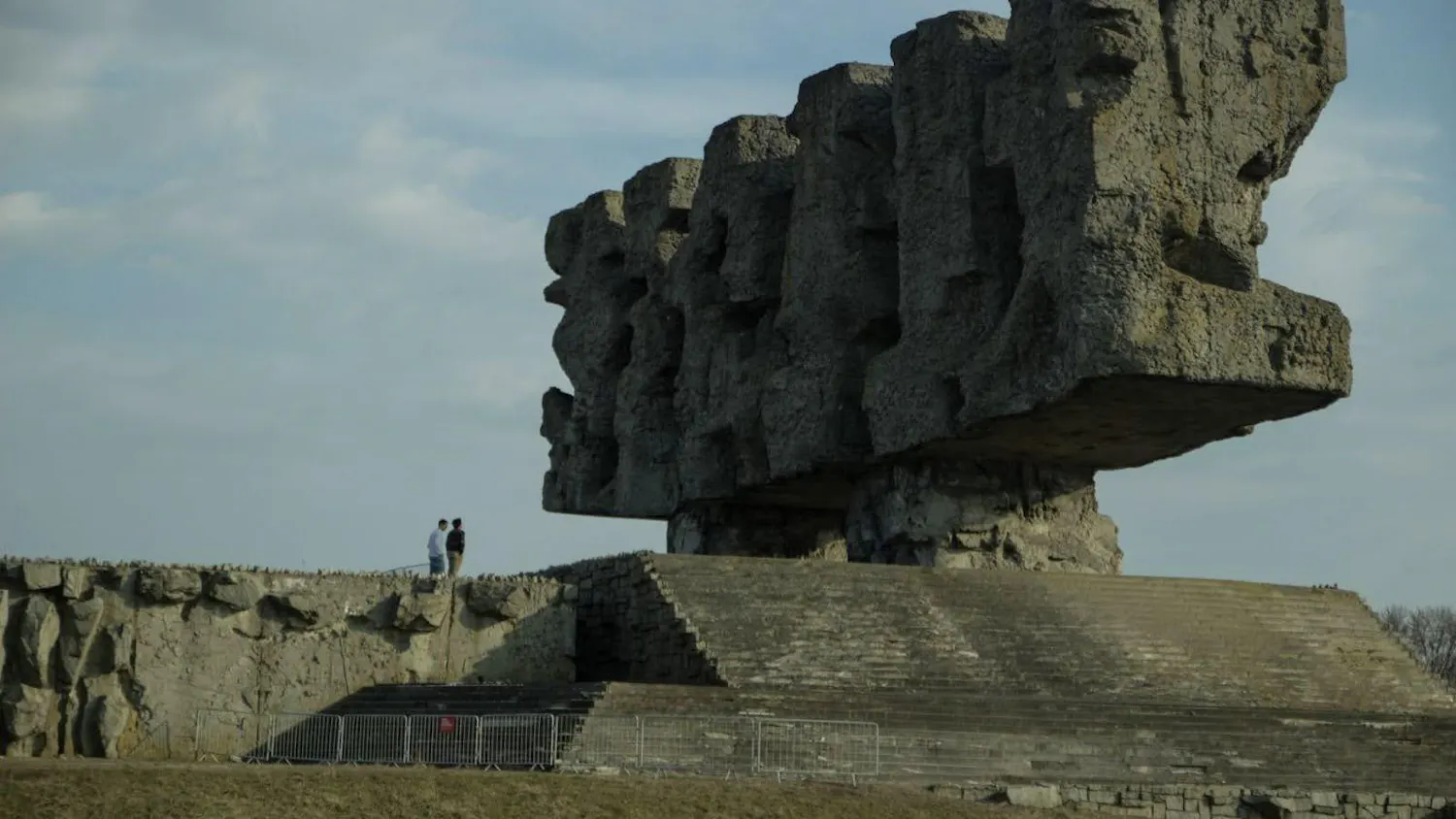 A photo showing a large, rectangular stone sculpture outside with stairs leading up to its platform. Two people are standing on the platform next to the sculpture.
