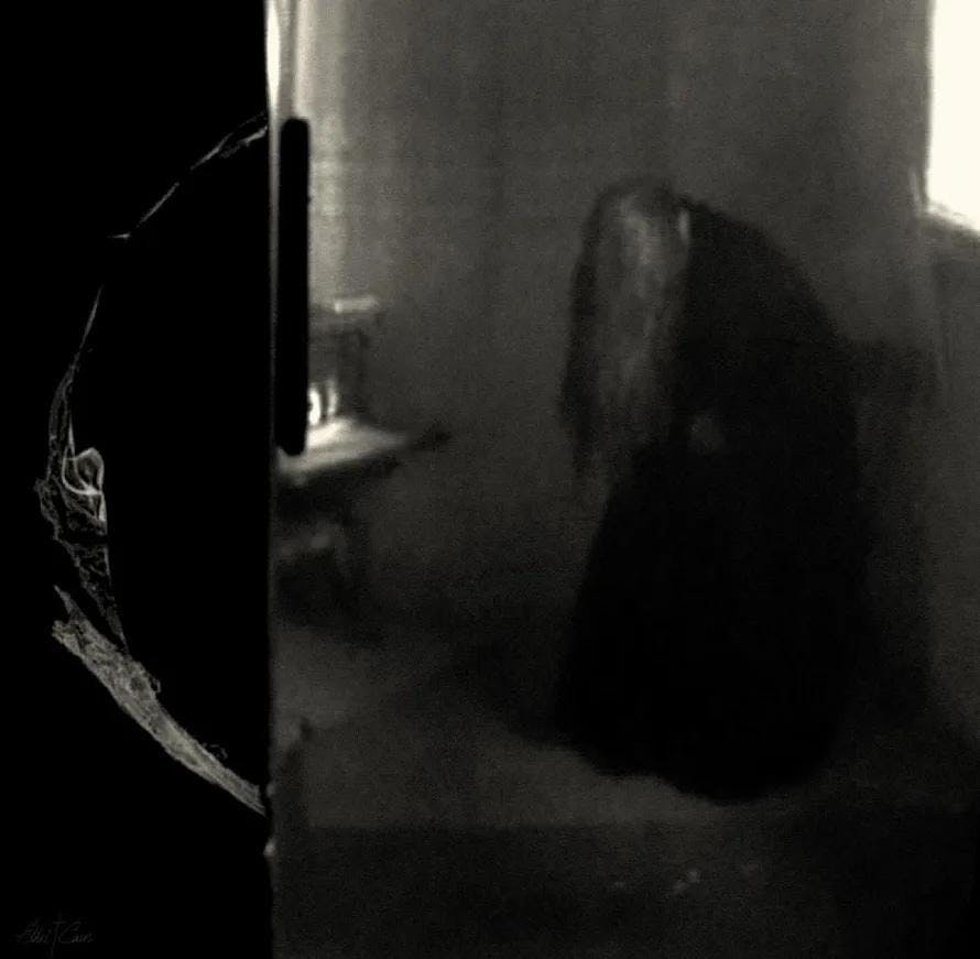 In the album cover for "Perverts" by Ethel Cain, in the background a long-haired girl in a long black dress sits on a chair with her head down and her hair over her face. The photo is black and white and the girl is blurry, and there is a sharp black rectangle in the foreground.