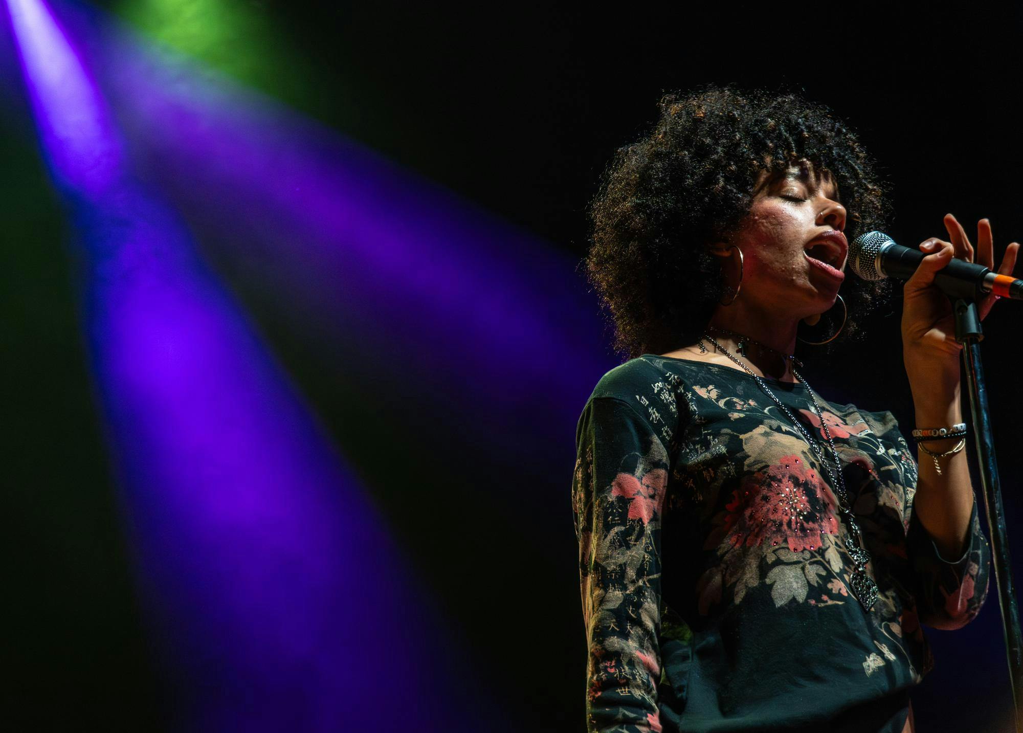 A woman with curly hair holds a microphone, illuminated by purple and green stage lights.