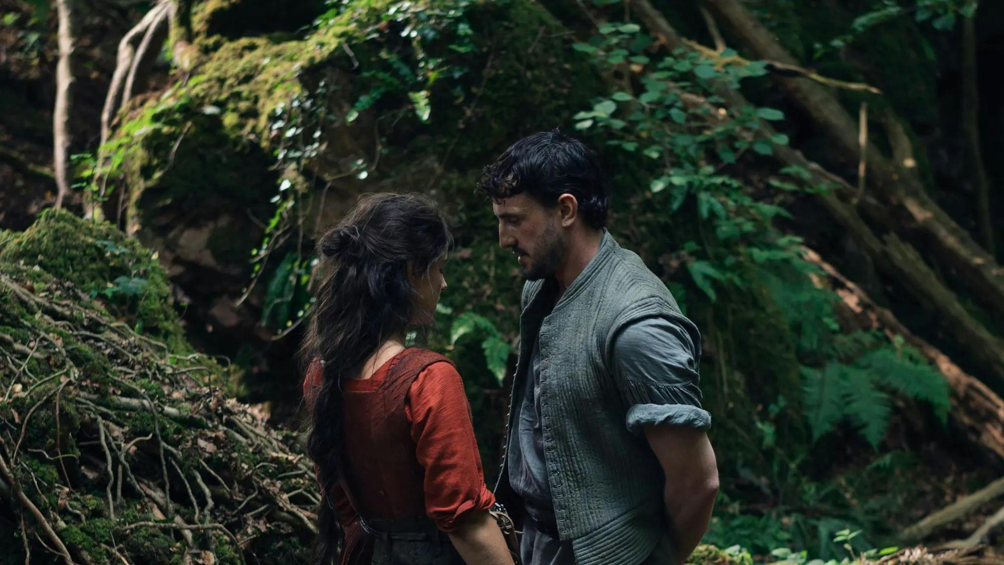 Still from the movie Hamnet, with William Shakespeare (Paul Mescal) in a gray open button up and Agnes Shakespeare (Jessie Buckley) in a red dress and loose brown hair facing each other in a lush green forest.