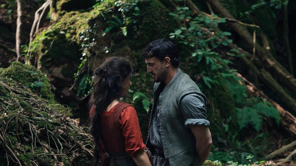 Still from the movie Hamnet, with William Shakespeare (Paul Mescal) in a gray open button up and Agnes Shakespeare (Jessie Buckley) in a red dress and loose brown hair facing each other in a lush green forest.