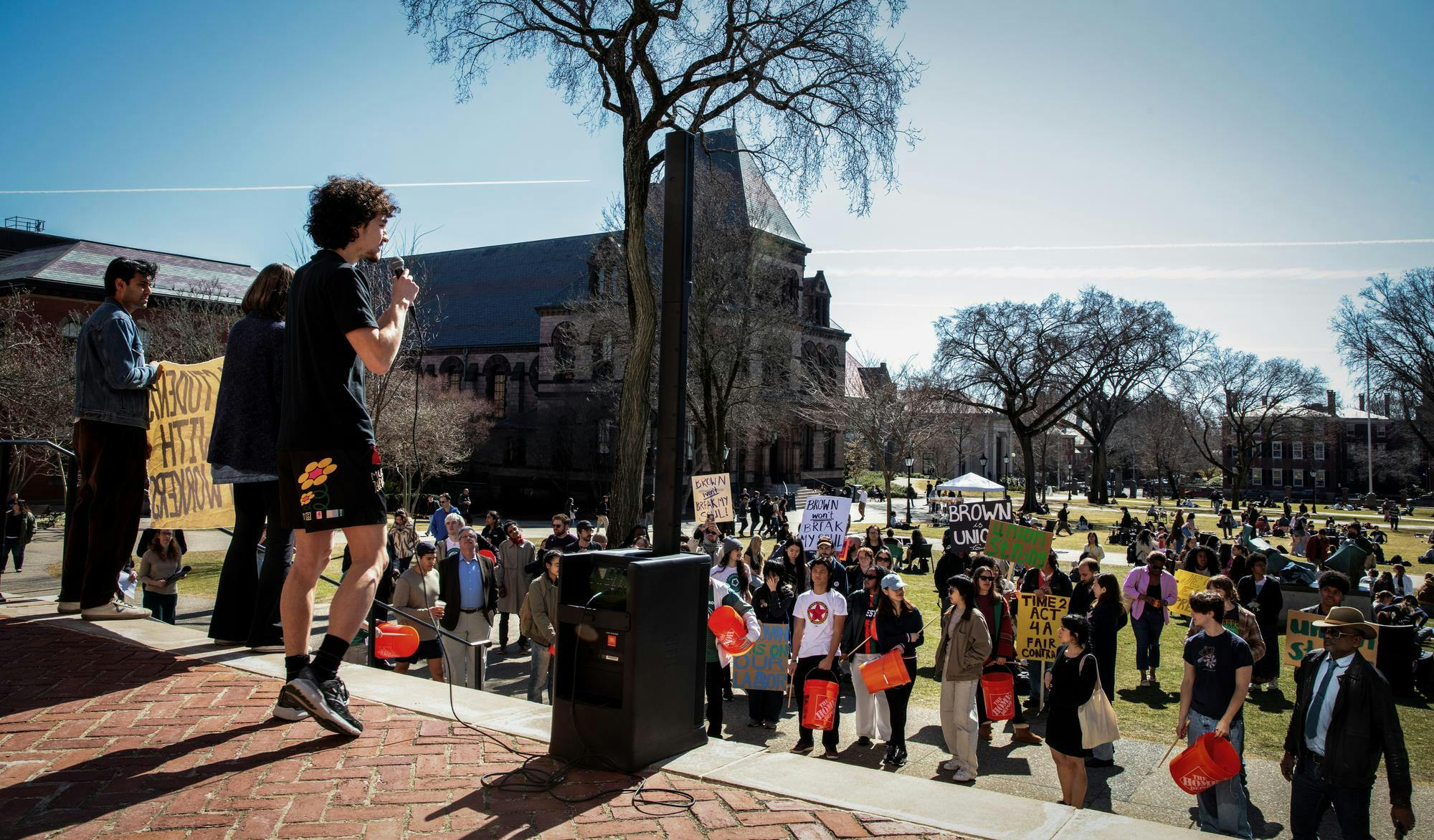 Photo of a man speaking at the top of stairs down to a crowd with a microphone and speaker, while various crowd participants hold signs and make-shift drums.