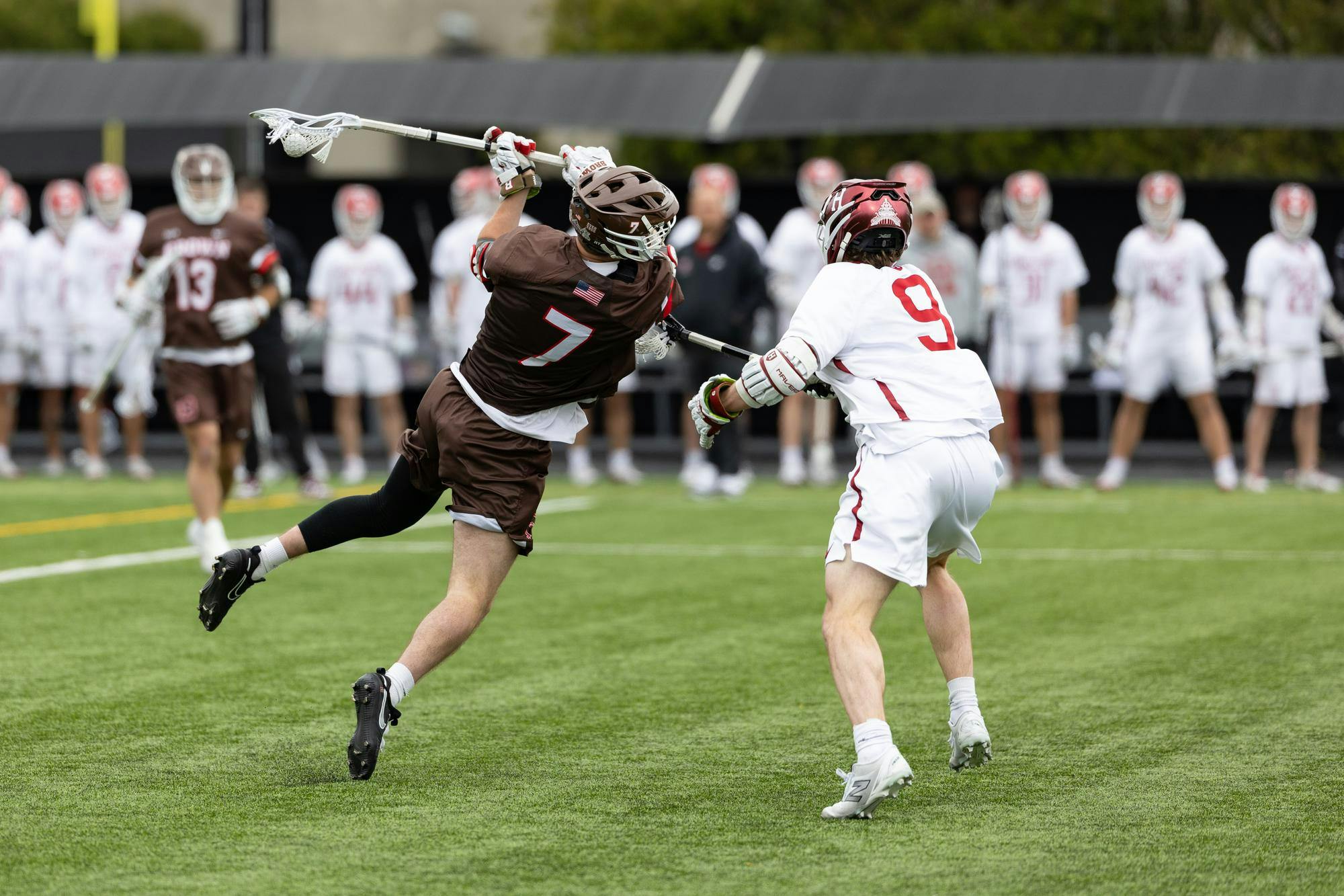 Action shot photo of a Brown lacrosse player mid-air against a Harvard player. 