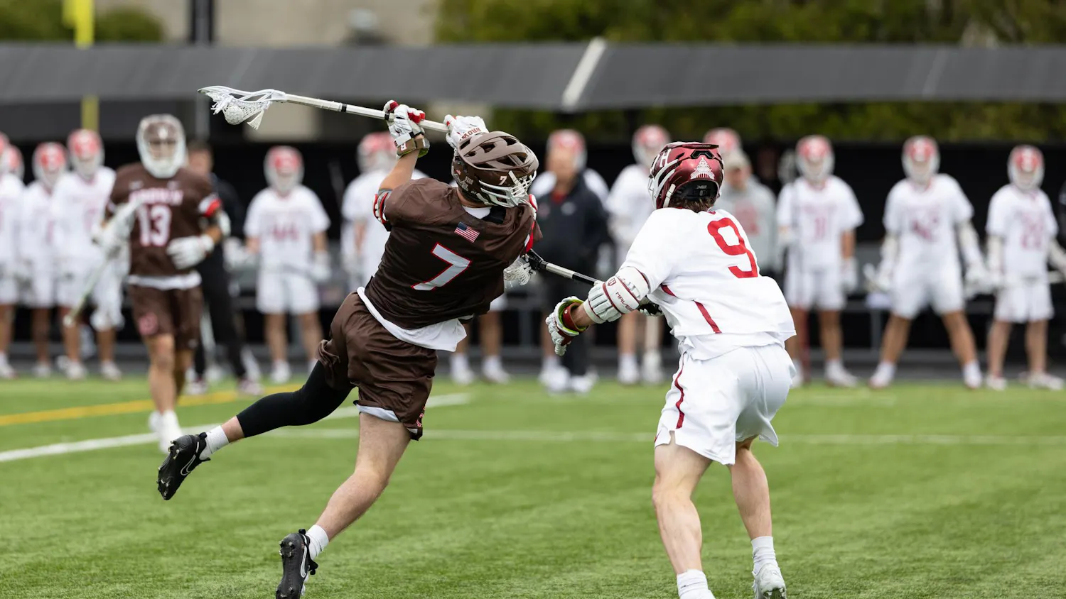 Action shot photo of a Brown lacrosse player mid-air against a Harvard player.
