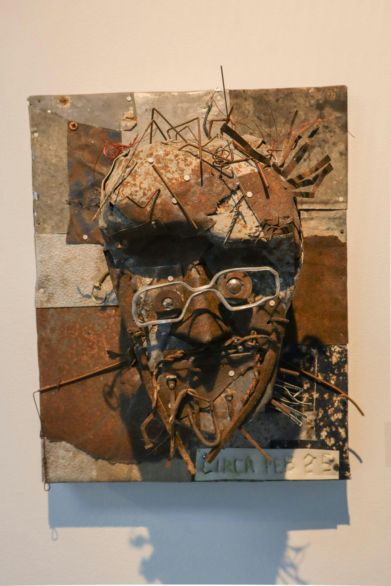 Photo of a sculptural mask made of discrete metal parts mounted on a wall.