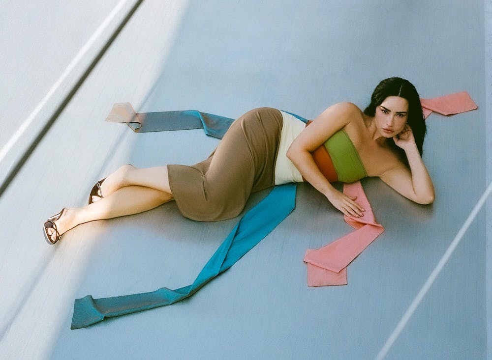 A woman laying on her side wearing a dress with a thick green, red, white, brown, pink, and blue stripe and brown heels.