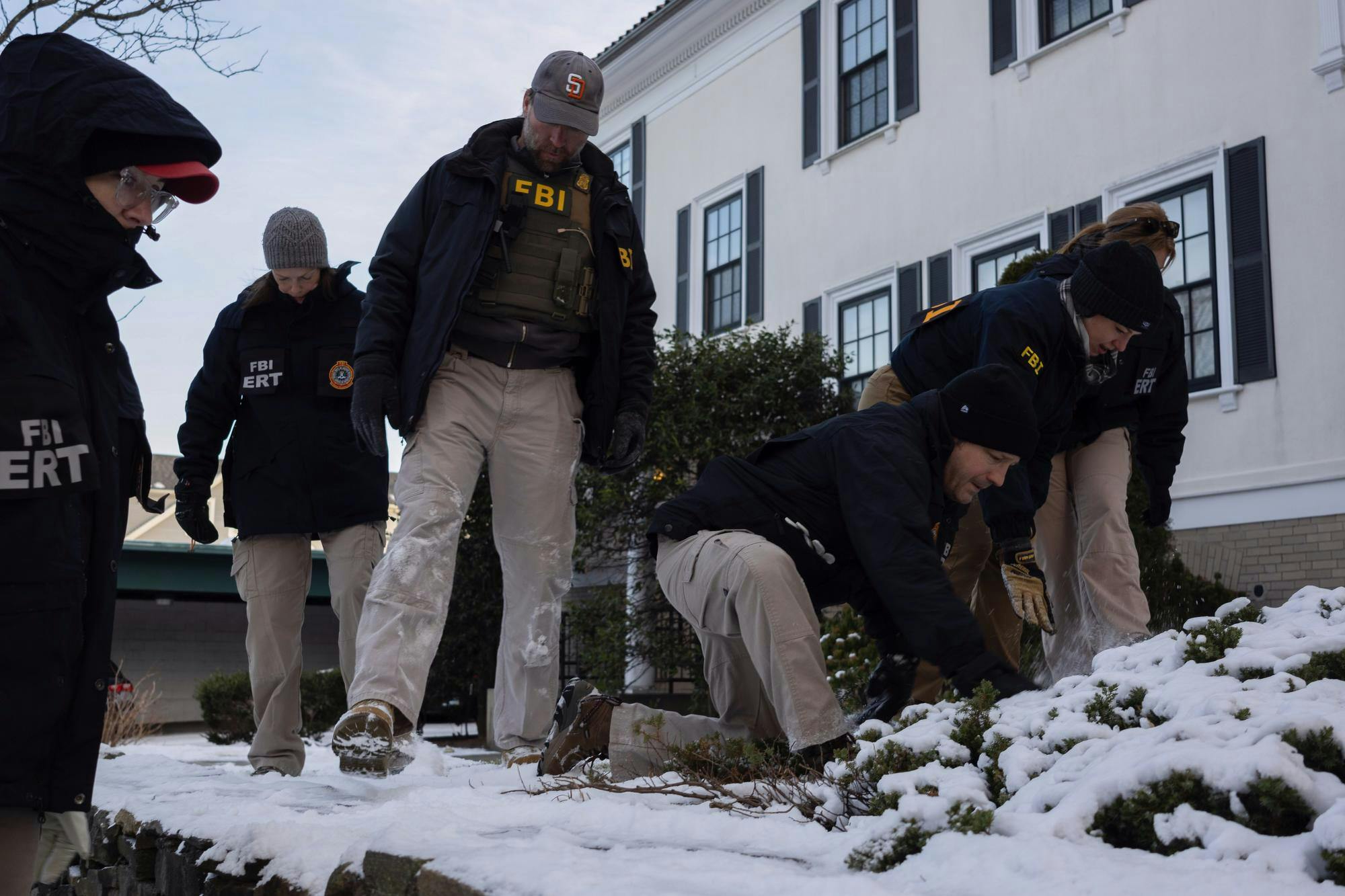 Photo of six FBI agents sweeping through snow with their feet in front of a white house, with some agents kneeling, brushing snow away from foliage with their hands.