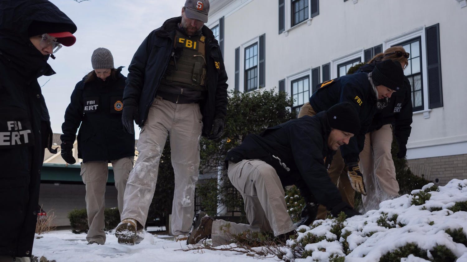 Photo of six FBI agents sweeping through snow with their feet in front of a white house, with some agents kneeling, brushing snow away from foliage with their hands.