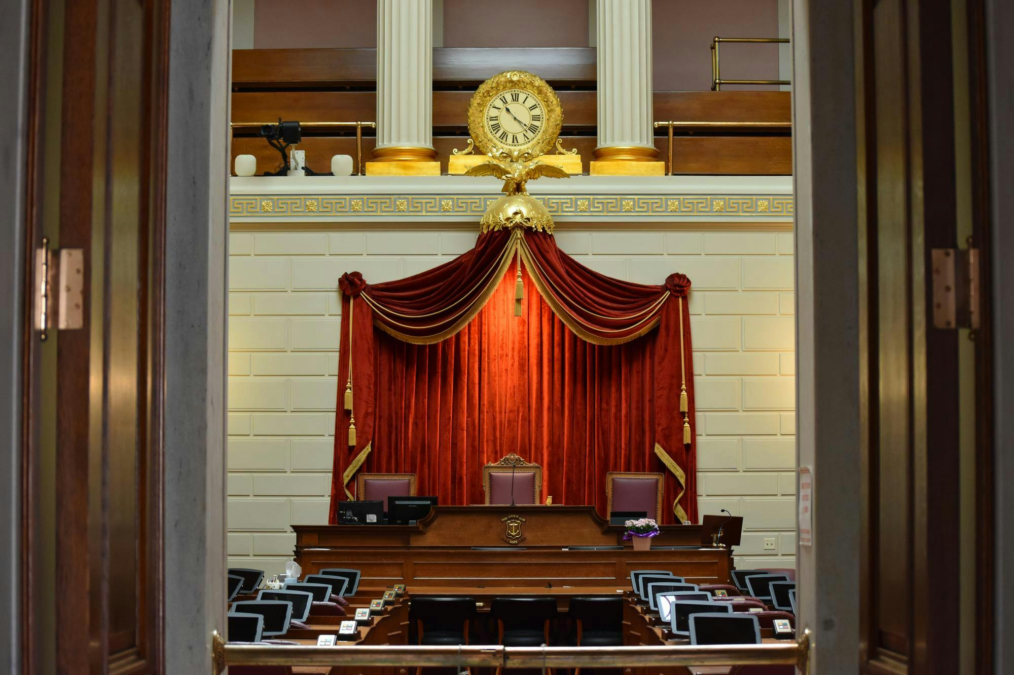 A photo of the interior of the Rhode Island legislature. The room features a gold clock over a red velvet curtain and three central leather chairs.