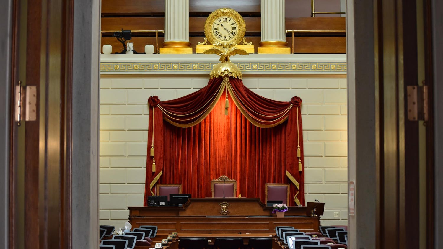 A photo of the interior of the Rhode Island legislature. The room features a gold clock over a red velvet curtain and three central leather chairs.