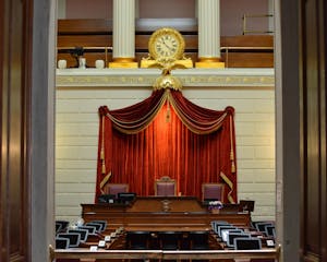 A photo of the interior of the Rhode Island legislature. The room features a gold clock over a red velvet curtain and three central leather chairs.