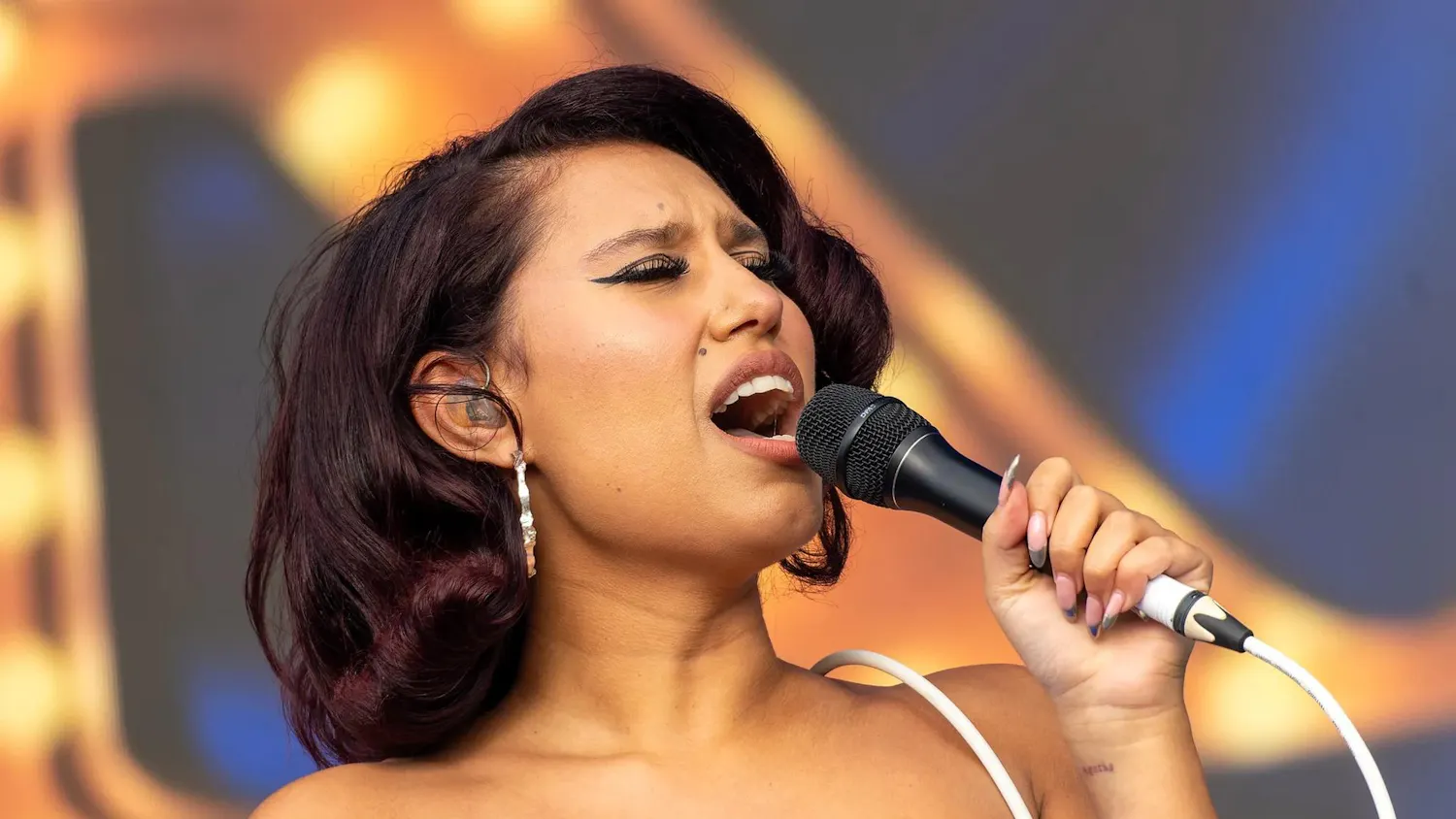 Photo of a woman in a blue strapless dress and silver hoop earrings singing into a microphone with her eyes closed and the microphone wire draped over her shoulder.