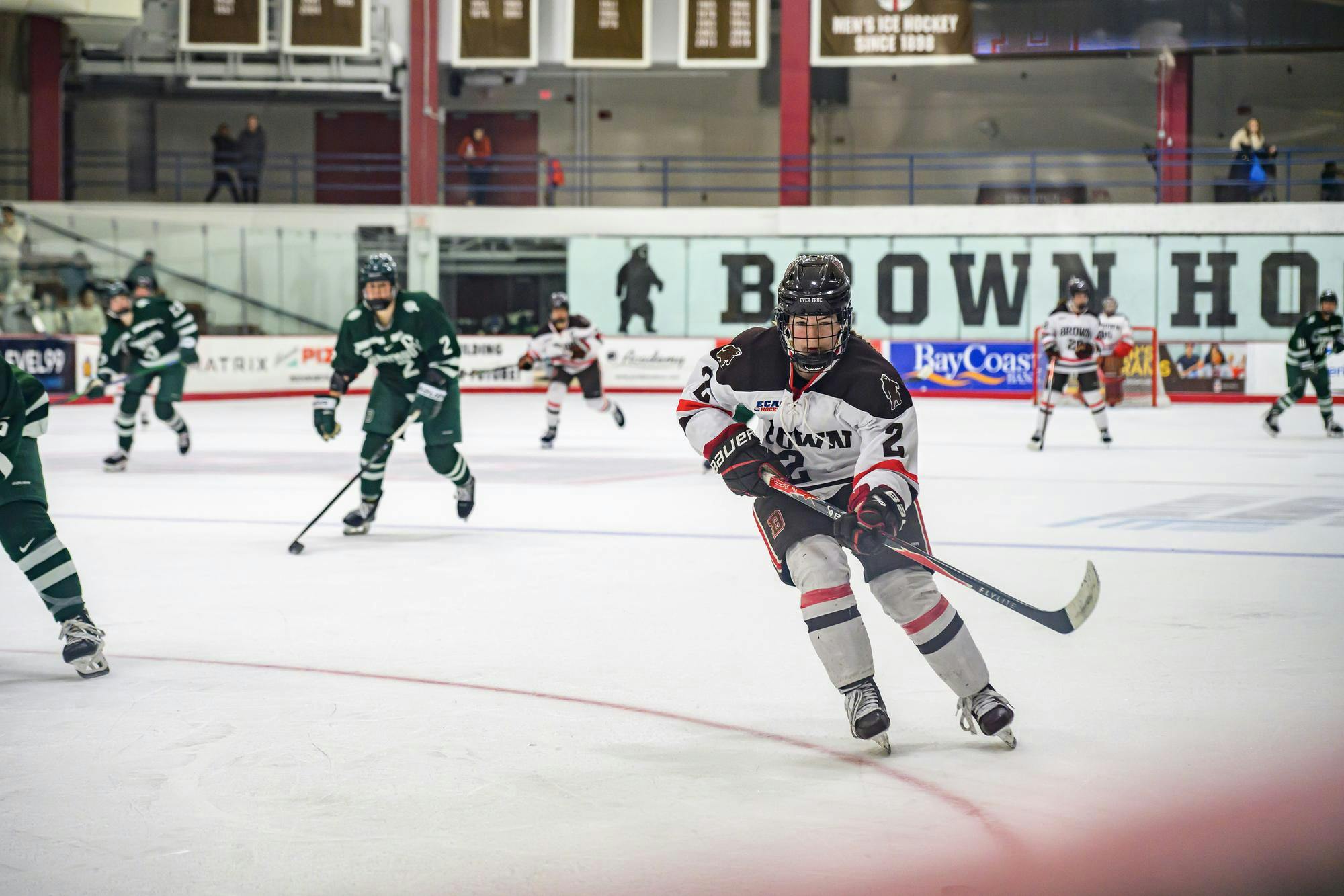 A photo of a Brown University hockey player holding their hockey stick while skating on ice towards a sliding puck with other players in the background. 